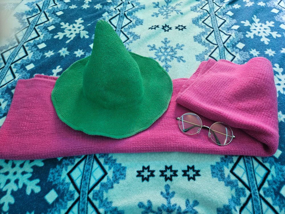 A trio of Cosplay items laying on a snowflake blanket. There's a green hat, large round pair of glasses, and a pink scarf. The Cosplay is Ralsei from the videogame Deltarune. By Toby Fox. Its quite good.