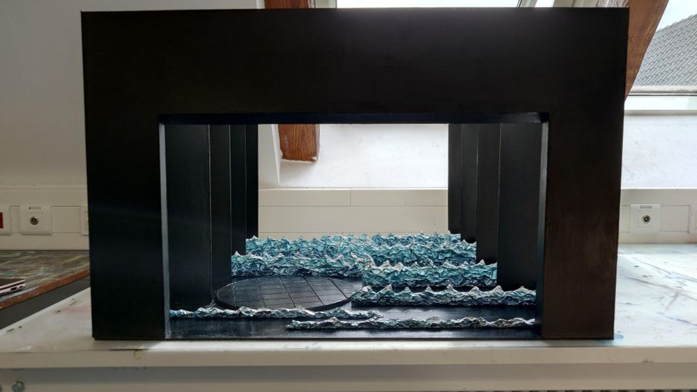 Front view of the current state of the stage set model. It is 68cm wide and 42cm tall. You can see the black stage, consisting of a stage portal, shiny floor and narrow side walls. And you can see the round black floor for the interior spaces, with large tiles. In addition, there are several theatre waves on the stage, which become larger towards the back, covered with aluminium foil and sprayed with metallic spray paint in a light-dark blue gradient.