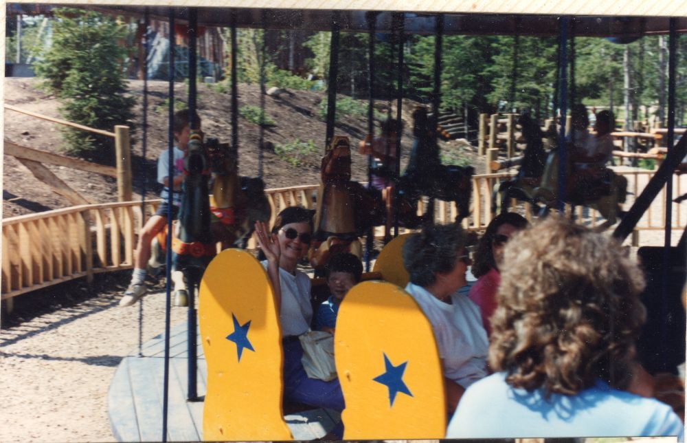 Merry go round with horses and benches, with wooden roller coaster in background. Benches are yellow with blue stars.