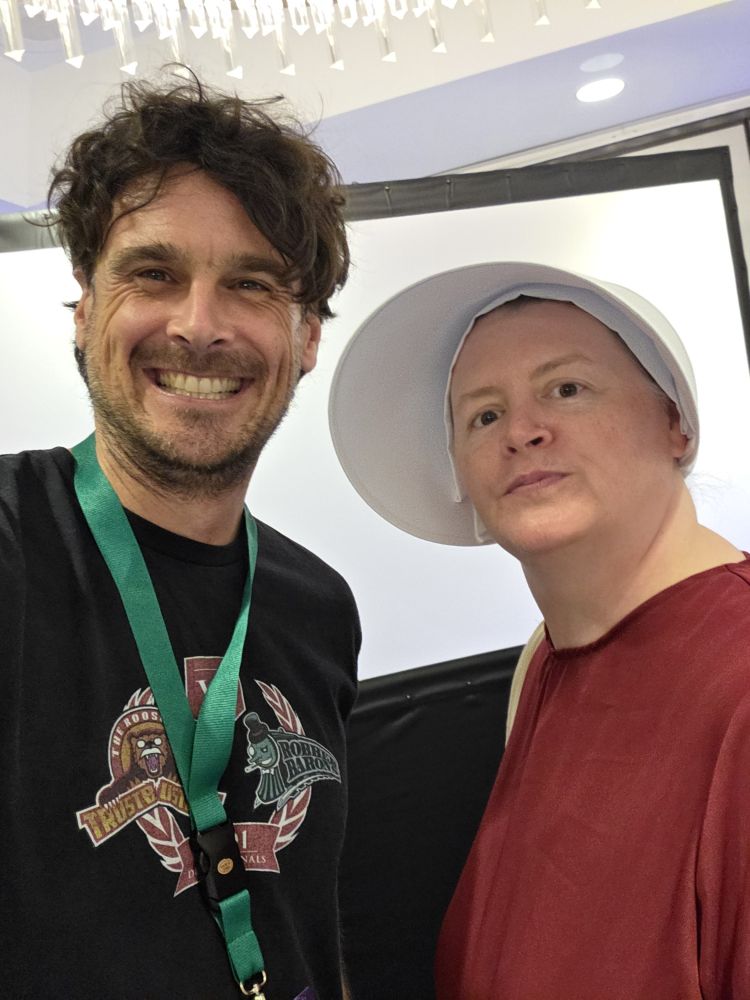 Selfie of Chris Kluwe smiling on the left, wearing a black shirt and green lanyard. Christine on the right in cosplay from "The Handmaid's Tale" wearing a red dress and white bonnet while trying to stay in character and not smile.