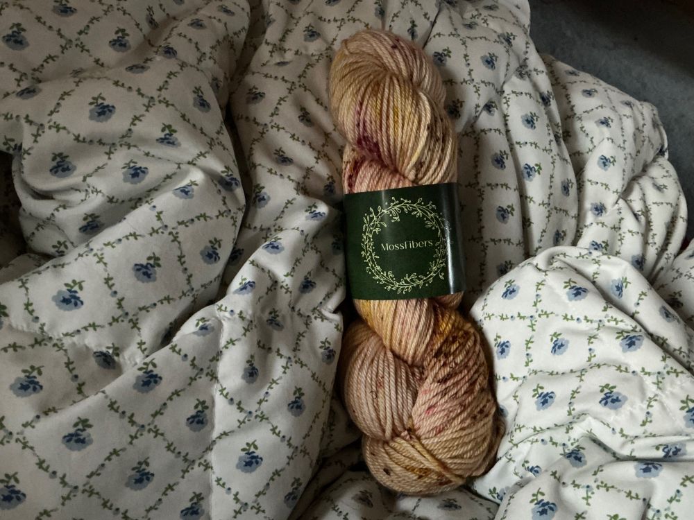 Yellow/orange-ish variegated and speckled skein of yarn on a white blanket with blue flowers.