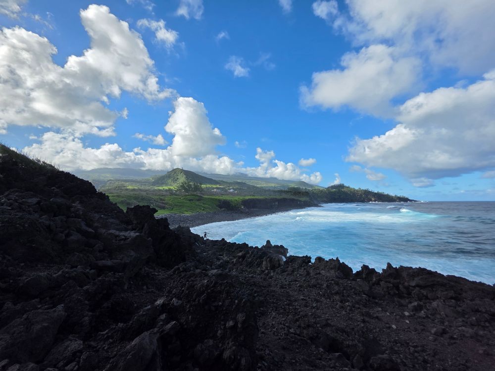 View from a rocky ground of Reunion island's south coast, the sky is sunny but with a fair amount of clouds, the ocean is on the right of the picture and the luxurious vegetation beyond the galet beach is on the left. All the foreground is an old lava field reduced in some  sharp rocks.

