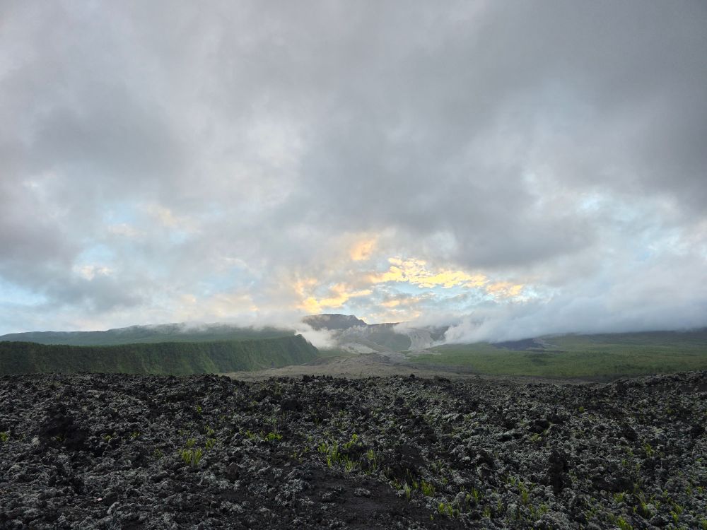 A view of Reunion island volcano from the south east coast, I'm taking the picture from one of the old lava flow, some vegetation has started to sprout from the lava rocks. On the far away is the volcano bathing in clouds and having a clearing sky just in the middle.