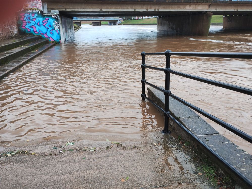 Brown river water ebbs at the bottom of a concentrate ramp. To the left steps leading up a bank. In the background a bridge over the river. The river is full and moving fast. There is foam and debris on it..