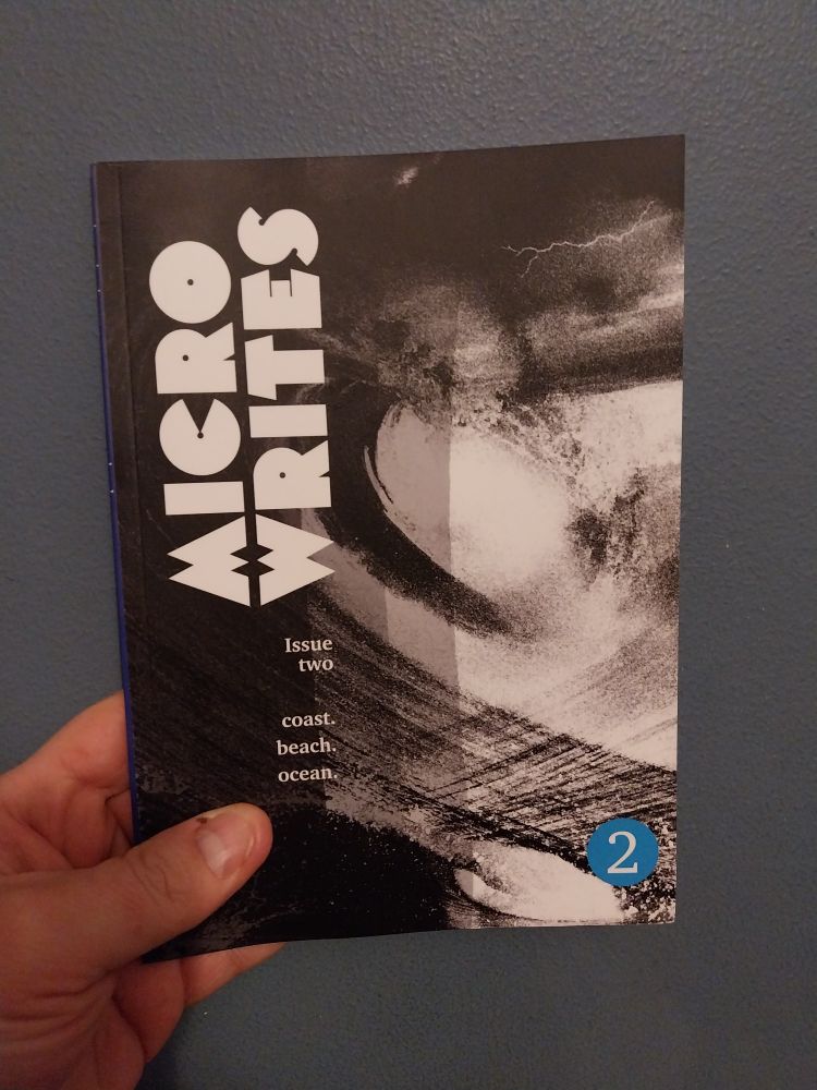 Microwrites issue 2, a black and white covered zine with the sub heading coast, beach, ocean held up to a dark blue wall
