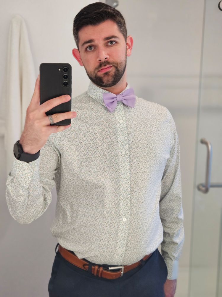 A hotel mirror selfie from the waist up, dressed in a button down shirt, slacks, and a bowtie.