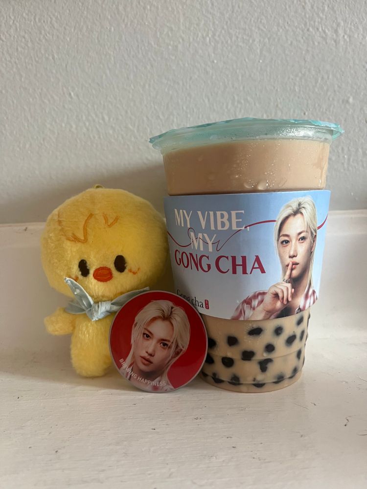 brown sugar milk tea with boba from gong cha with a felix cup sleeve saying "my vibe my gong cha" and has his face on it and a little pin that also has his face and a bbokari from the stray kids pilot merch