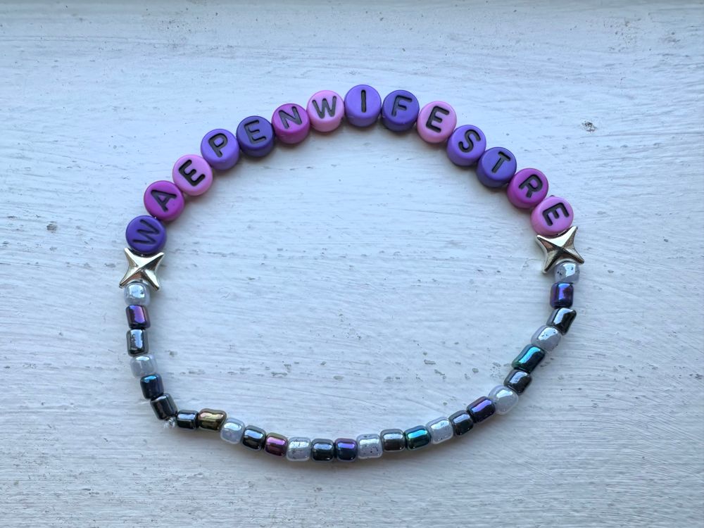 a handmade beaded bracelet in a purple-pink-silver-ish colour scheme that says “waepen wifestre” with silver sparkles on either side of the word 