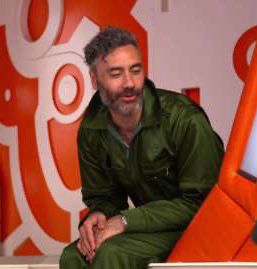 Taika Waititi, 41 years old. Wearing his olive green jumpsuit, during the filming of Thor: Ragnarok. He is sitting with his hands in his lap speaking to someone offscreen. 
