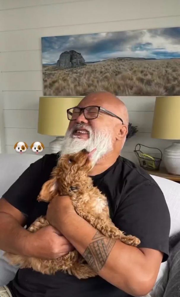 Dave Fane is holding a little curly dog as it tries to lick his face. Dave is laughing with his eyes closed as this is happening because obviously he was created from nothing but pure joy and abundant sunshine. There are two dog emojis in the corner. 