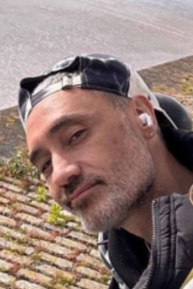 Sound the alarm, it’s fuckboi Taika Waititi. In what may be the most damaging-to-me photo in existence, Taika is sitting, looking back at the camera with a smirk? But like a 1993 B-boy smirk? His hat is on backwards and he’s wearing a coat whose collar is popped up right under his construction-grade angled jawline. So sharp it can be used in a street fight. The presence of an earbud only draws our eye to the diamond stud in his ear. I remain awash. 