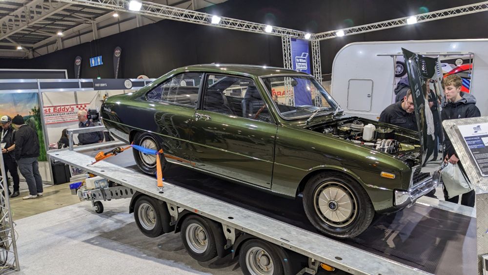 A dark green Mazda R100 Coupé looks down on the humans from atop a trailer, its forward-hinged bonnet open.