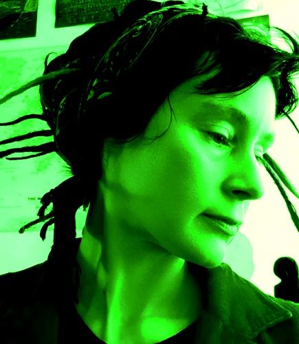 A photo of Penny Blake with her head to one side showing dreadlocks tied up in a rose-patterned dikhlo. Penny is wearing a t shirt and a corduroy jacket. The whole image is tinted bright green.