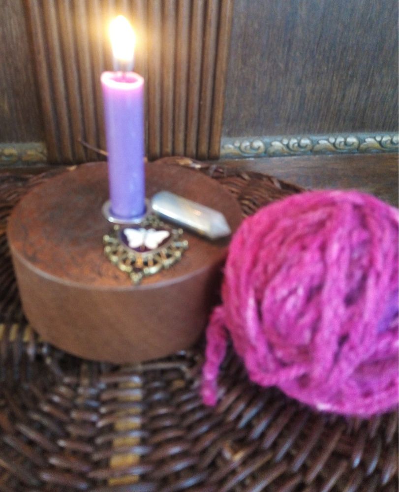A purple candle symbolising both mourning and protection is in a wooden candle holder with arcane engravings on it. The candle is on a hand woven willow alter with a wooden backing. Placed around is a brass and enamel butterfly charm symbol of new life and freedom and crossing the veil between worlds, a tricksy little crystal that has a mind of its own and will not be told what to do, and a ball of hand woven yarn for kitty boy Jack to play with in a joyful pink colour in celebration of his life and the love he brought and shared.