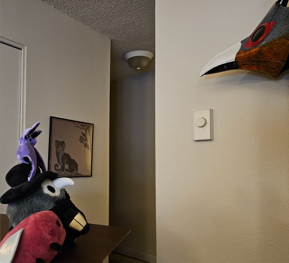 An adorable plush bat, a rotund plush plague doctor, and eggbug all ponder a painted plague doctor mask hanging above them. An anatomically questionable tiger is framed in the background.
