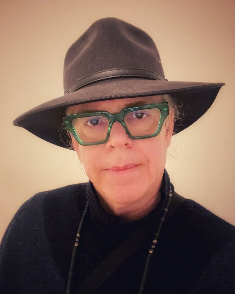 head and shoulders portrait of a man with thick green crystal frame glasses, black sweater and gray felt broad brim hat…