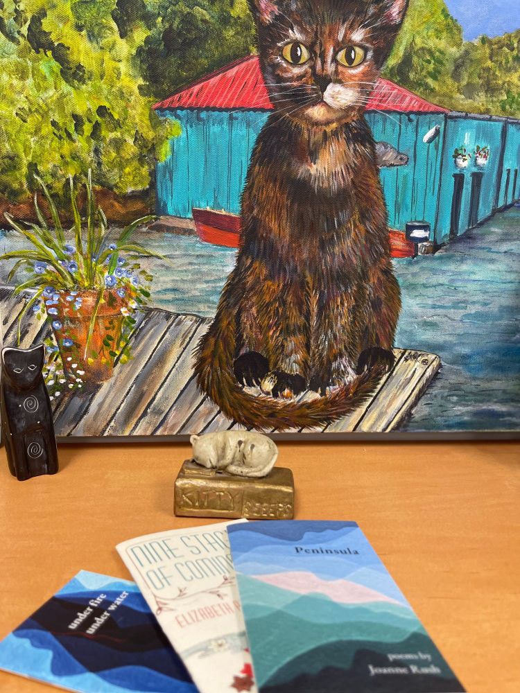 Foreground: Three chapbooks published by tiny wren (under fire | under water; Nine Stages of Coming Out; and Peninsula

Middleground: A folkart carving of a sleeping cat on a book

Background: Painting of a cat sitting on a table and next to a terracotta pot of flowers; behind the cat, turquoise blue building with a red roof
