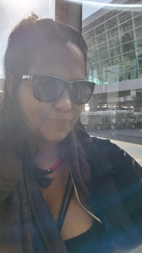 Indigenous woman in her forties (looking thirties), brown skin, black hair, wearing sunglasses, black shirt and cardigan, and a coral snake necklace. Smiling like Monalisa (give me a break, I'm describing myself here).
