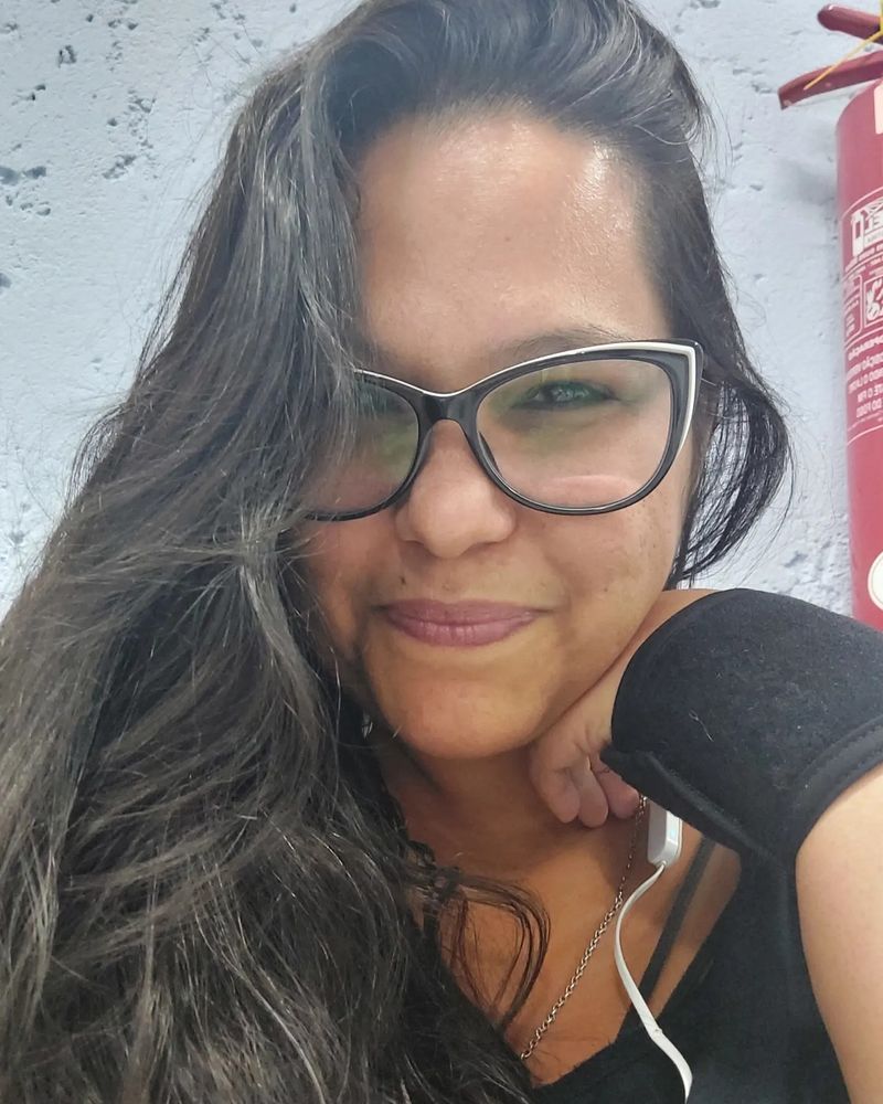 Smiling Mestiza/Indigenous woman with brown skin, black hair, glasses, in her forties (looking thirties. I'm writing my own description, give me a break, ok? Lol). Her hand is under her chin, and she's wearing a wrist brace.
