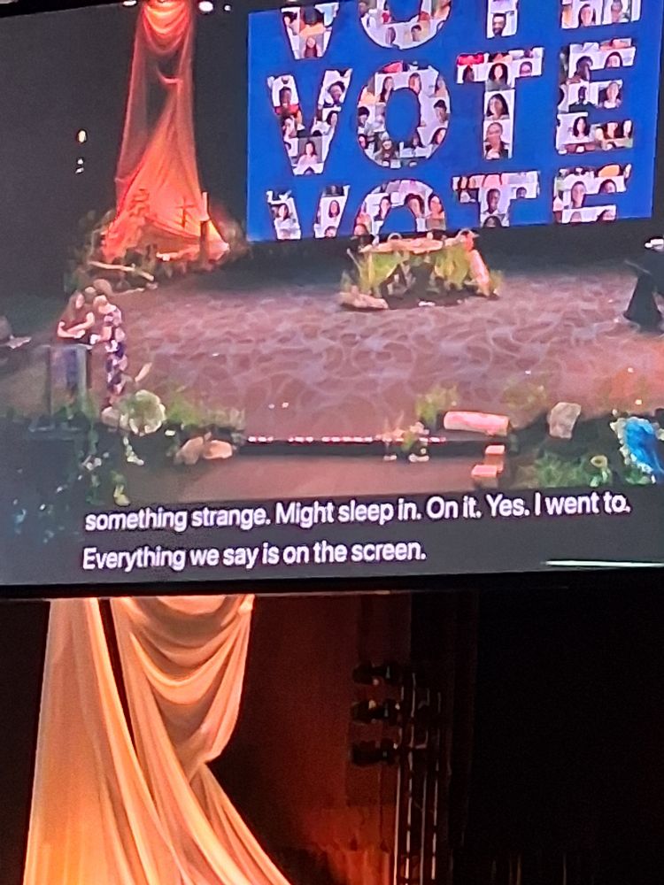 Photo of large screen during voting drama with closed caption "Everything we say is on the screen."
