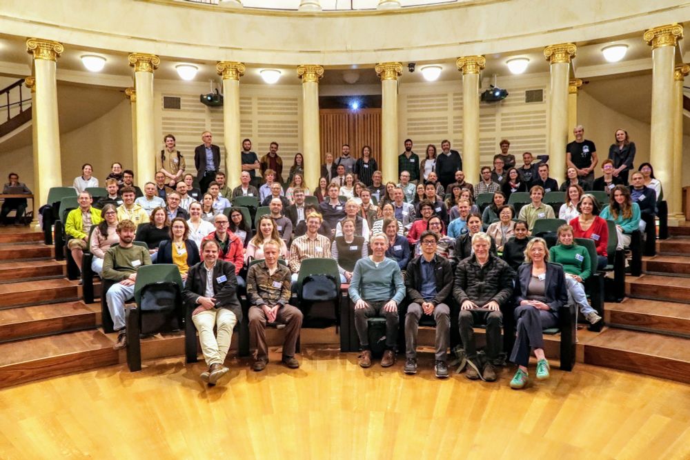 A group photo of the attendees of the Liège Colloquium, sitting in rows. Wooden floor in the front and columns in the back.