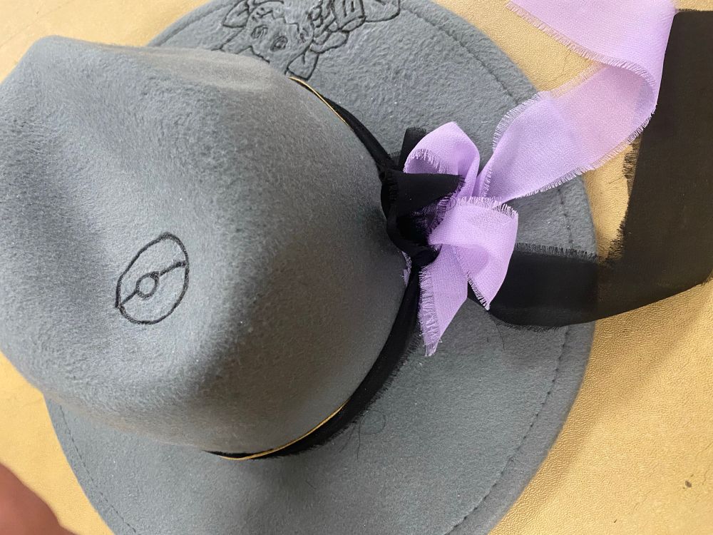 a grey felt hat with a pokeball image burnt into the crown. there is a black and purple bow on the back, the tails of the bow hanging over the side of the brim.