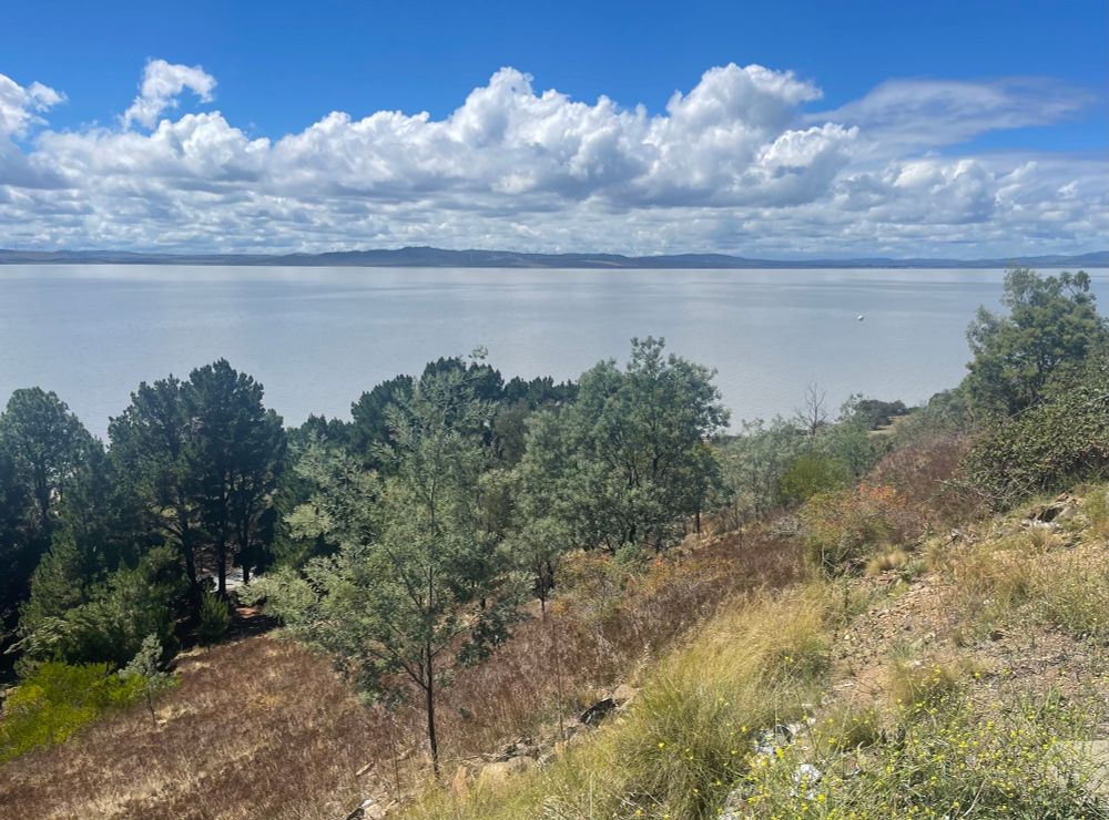 A scrubby hillside, a large lake under a blue sky and line of clouds