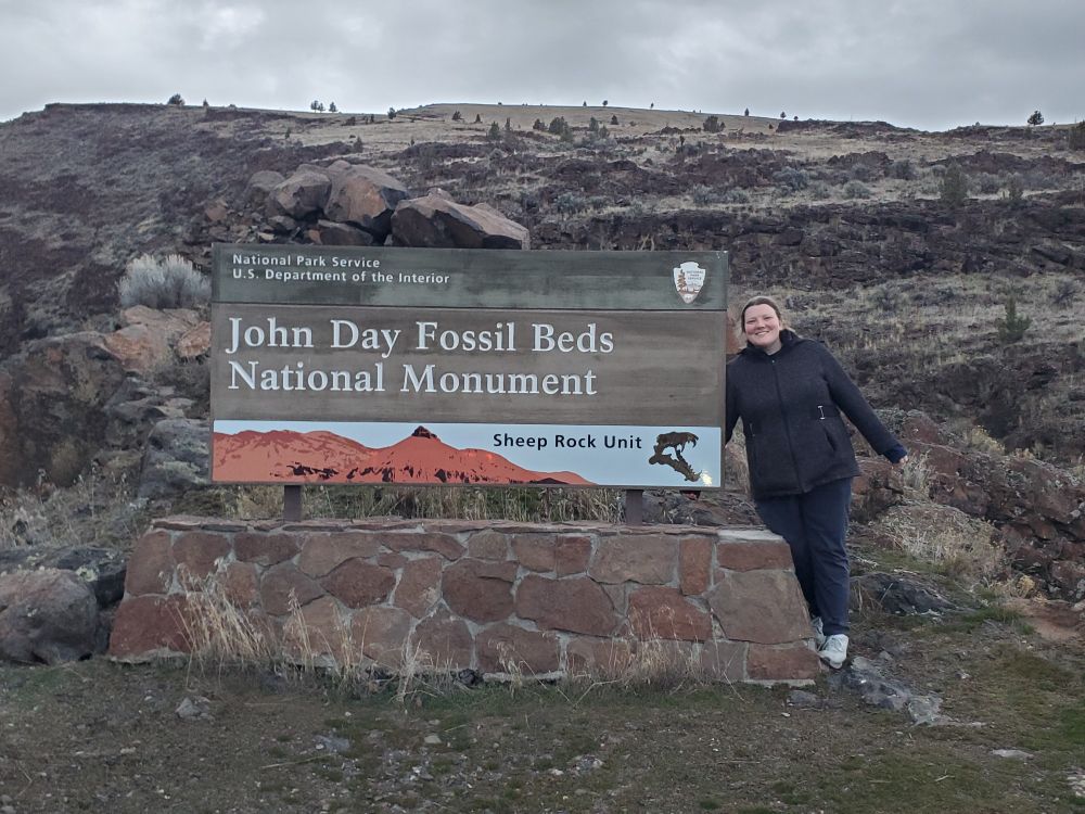 Woman standing next to park entrance sign. Sign says, John Day Fossil Beds Narional Monument, Sheep Rock Unit.