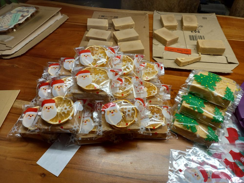 Photo of a table with soap bars. Some are packed in bags with Christmas motifs like Santa Clause or Christmas trees. There are over 20 soaps on the table.