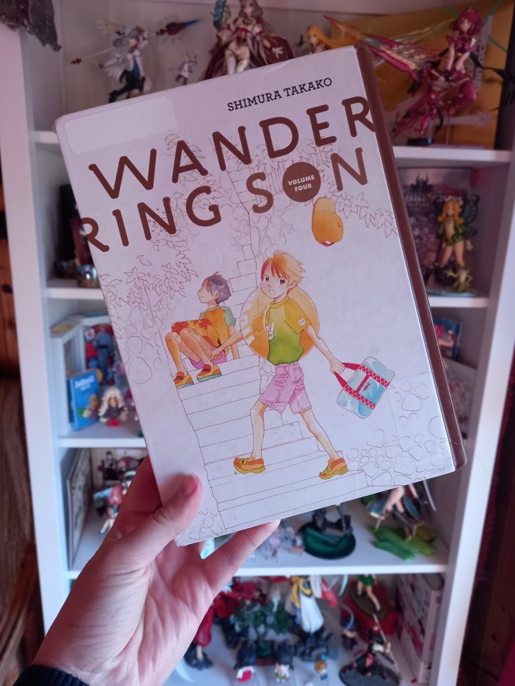 A hand is holding the 4th volume of the manga "Wandering Son". It shows the protagonist Shu making a step forward, looking at the reader while holding a bag in their left and and a bathing ring over their other shoulderm in the background Takatsuki sits on the stairs and looks at the sky smiling.