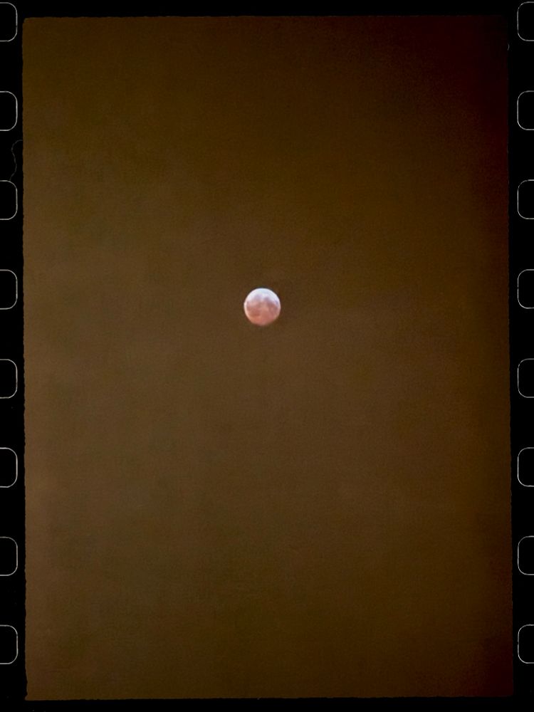 A photo of the moon using a phone camera. The moon is small but slightly red and centered. Some 35 mm film sprockets are along the side.
