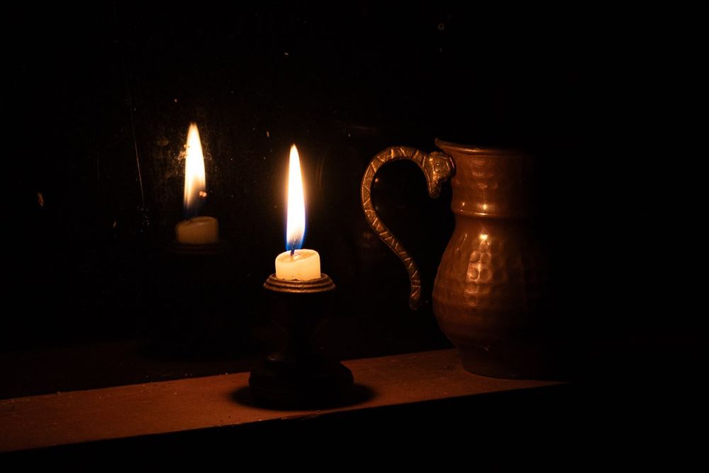 Foto colorida noturna de uma vela acesa em um pequeno castiçal refletida no vidro de uma janela. No lado direito um pequeno jarro.
O fundo é predominantemente escuro.