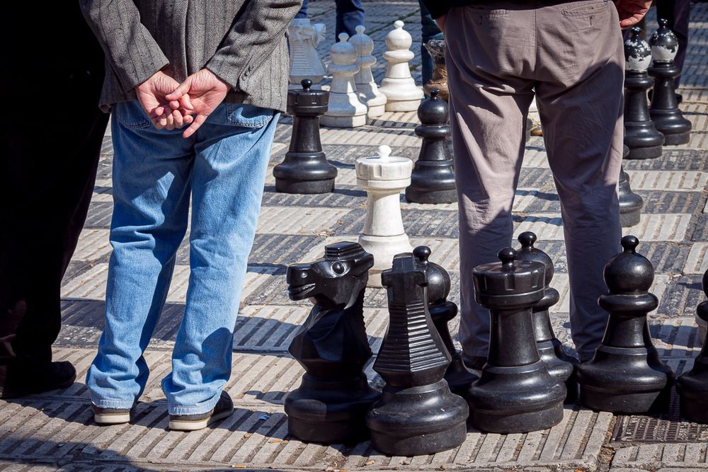 Foto colorida de um jogo de xadrez em tamanho grande, em uma praça com pessoas de costas, visíveis da cintura para baixo, observando as peças no tabuleiro. As peças são brancas e pretas assim como o tabuleiro pintado no chão.