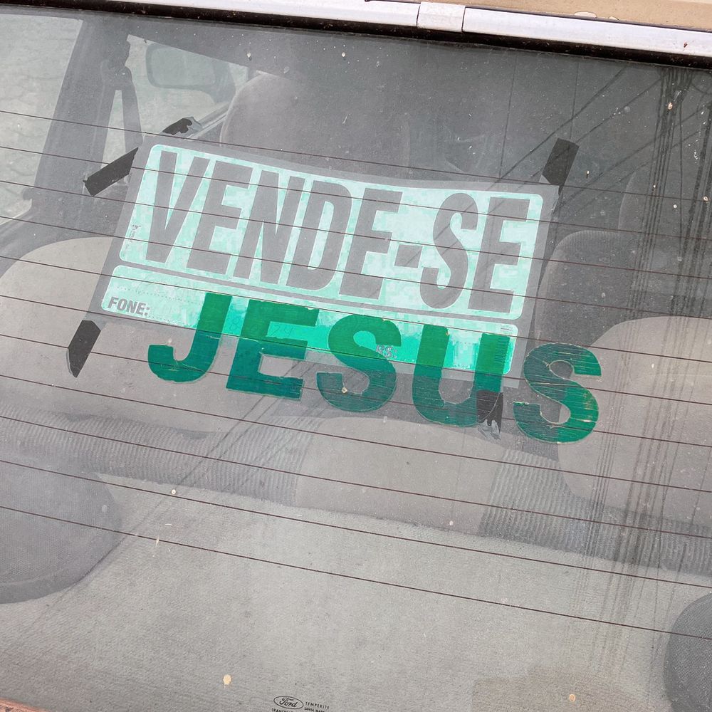 Foto colorida de uma placa de ”vende-se" colada no vidro de trás de um carro. Abaixo da placa um outro adesivo onde está escrito ”jesus".