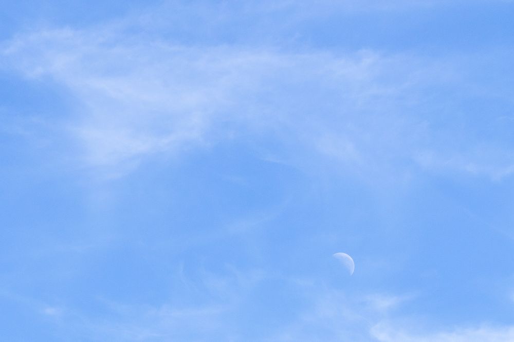 Foto colorida do céu azul com algumas nuvens brancas e a lua minguante na parte inferior direita da imagem.
