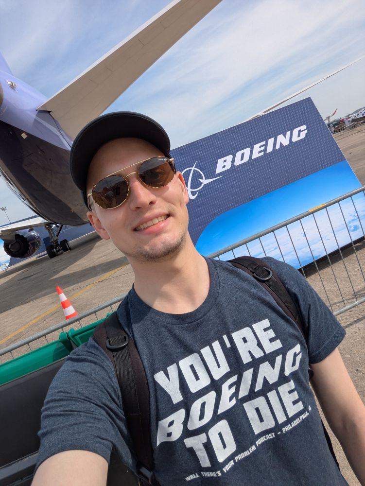 Selfie of me standing in front of a Boring 777 and a Boeing logo, smiling, wearing a hat, sunglasses and a T-shirt which reads "you're Boeing to die"