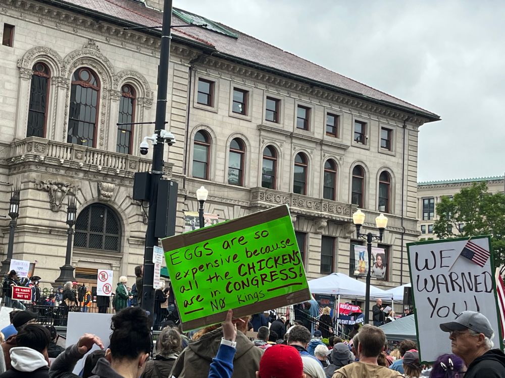 A crowd in the plaza behind Worcester (MA) City Hall, with a neon-green sign in the center reading: "Eggs are so expensive because all the chickens are in Congress! No Kings"