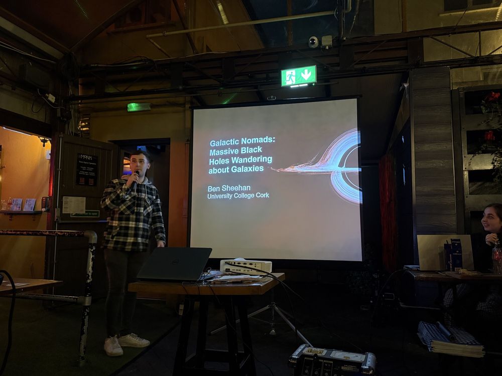 UCC Masters student Ben Sheehan gives a talk about massive black holes wandering in galaxies, pictured here standing by a projector screen featuring the iconic Interstellar black hole image