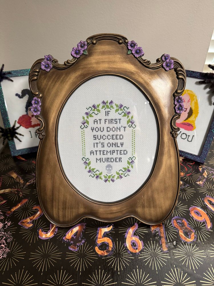 A frame with purple flowers around around the top has a hand stitched cross stitch that reads “if at first you don’t succeed it’s only attempted murder”