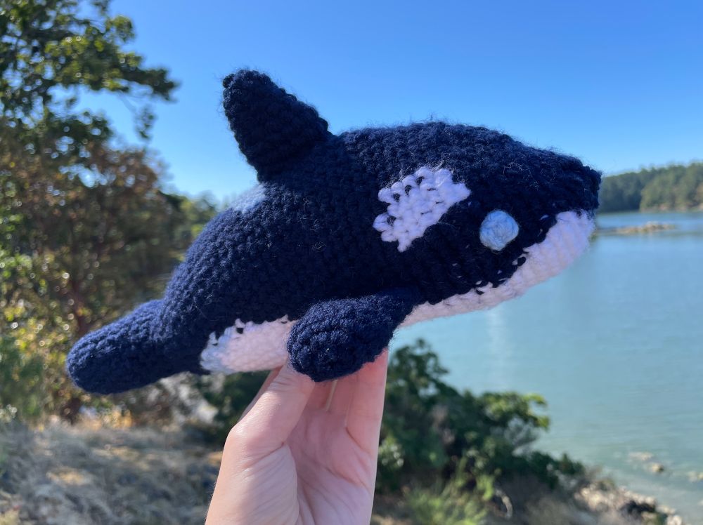a crochet orca, in profile, in front of a sunny inlet.