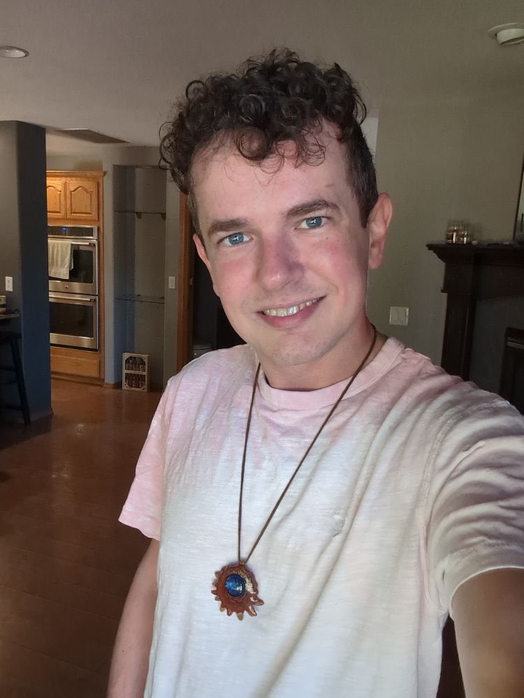 A selfie picture that I took the other day at home.

My general look: I have curly dark brown hair which I like to allow to grow out, while trimming my hair shorter on the sides and back. I have blue eyes and a pale complexion.

In this photo I am smiling at the camera and wearing a pink t-shirt and a necklace which has a resin-coated cross section from a pinecone and a blue glass bead on the front.