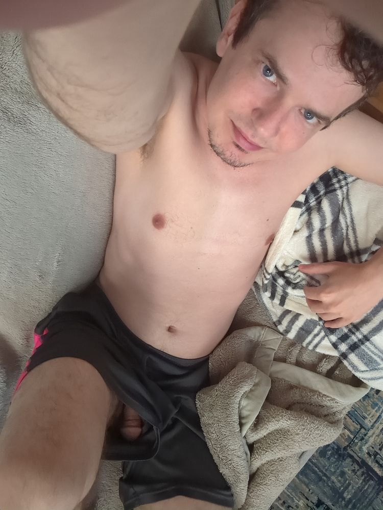 A selfie picture of me lying down on a couch, on top of a light green blanket, wearing nothing but black silky gym shorts with a pink stripe on the side. My face is looking up at the camera, and with one of my knees up, you can see up a leg of my shorts and clearly see the shaft and head of my flaccid penis.