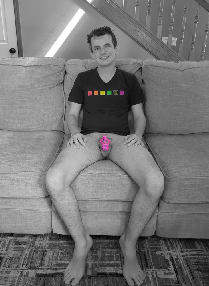 A mostly grayscale photo of me sitting on a couch in my living room, my face grinning at the camera, wearing nothing but a black t-shirt with gay pride colors on it. Below the t-shirt, I am bottomless wearing nothing but a pink chastity cage on my flaccid cock. While the photo is mostly in black and white, the gay pride colors and pink chastity cage are in their original colors.