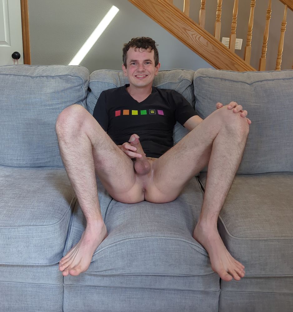 A photo of me sitting bottomless on top of a grey sofa, wearing nothing but a huge grin on my face and a black t-shirt with gay pride colors on it. My feet are both up on the sofa, legs spread apart, exposing the full bottom half of my body with my rock hard cock in my hand and my smooth shaved taint and boy hole all on clear display.
