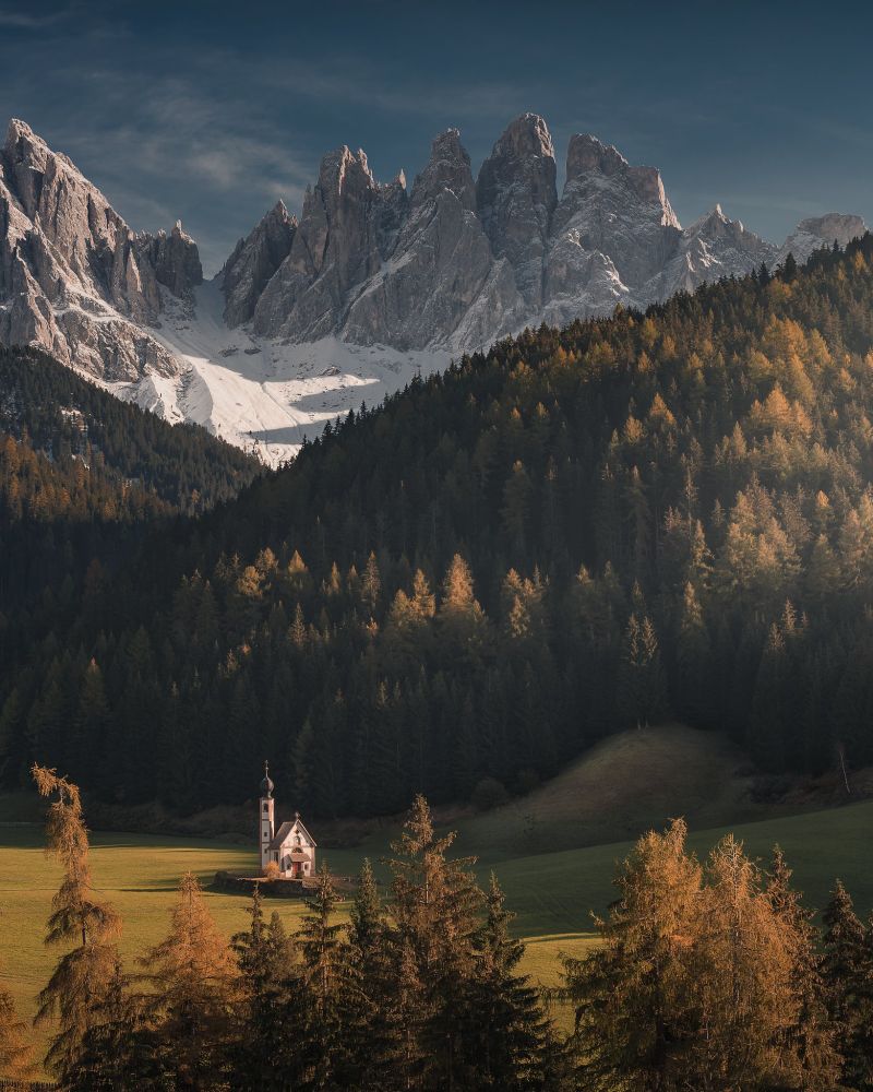 A picturesque mountain landscape with dramatic jagged peaks in the background, partially covered in snow, under a deep blue sky. In the midground, dense evergreen forests blanket the slopes, transitioning into a sunlit clearing in the foreground. Nestled in this clearing is a small, charming chapel with a tall bell tower, surrounded by green meadows and scattered autumn-colored trees. The scene conveys tranquility and isolation amidst nature’s grandeur.