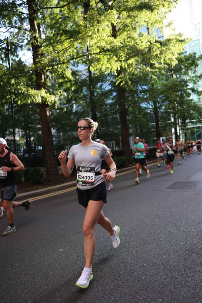 A woman is running in a competitive race along a road, wearing shorts, a grey t-shirt and sunglasses.