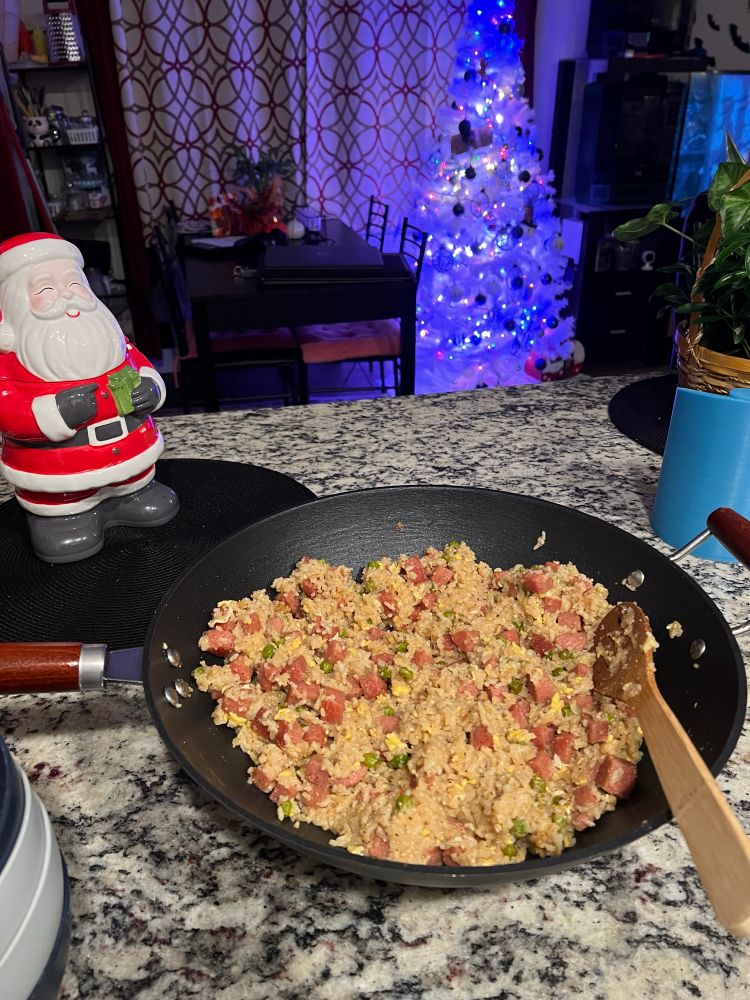 Pictured is a gray granite kitchen counter with a cast iron wok on top filled with fried rice, egg, spam, and green peas. In the background is a Santa cookie jar and a white and blue Christmas tree. 