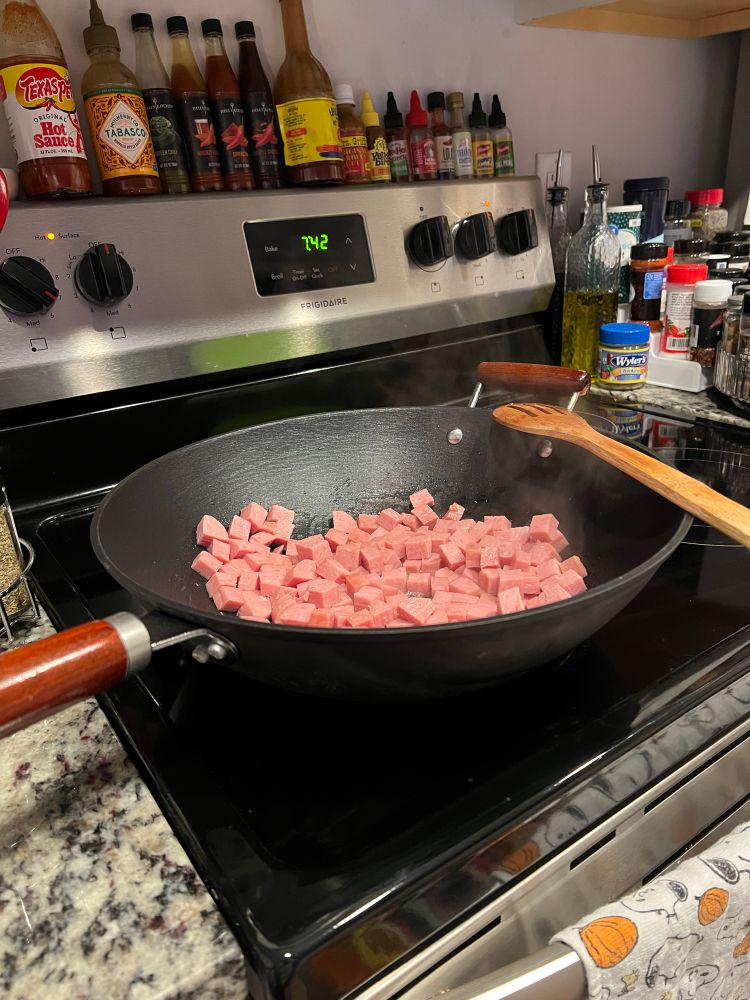 Pictured is a large cast iron wok on top of a black and silver stove top. Inside the wok is chunks of diced spam with a wooden spoon rested on the edge. In the background is lined up hot sauce bottles and seasonings. 