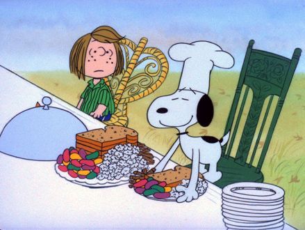 Chef Snoopy is setting down a plate of bread, jellybeans, and popcorn on a long white tablecloth table with a platter of said food and a stack of plates. Chef Snoopy is standing in a green chair beside of Peppermint Patty who is sitting in a gold colored chair. 