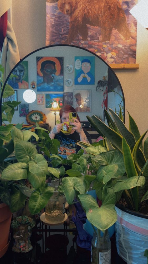 Pictured in the distant middle is me with no makeup in my pjs holding up a yellow Buccees coffee mug and yellow iPhone case. I’m taking a selfie in dome shaped mirror surrounded by lots of green plants. 